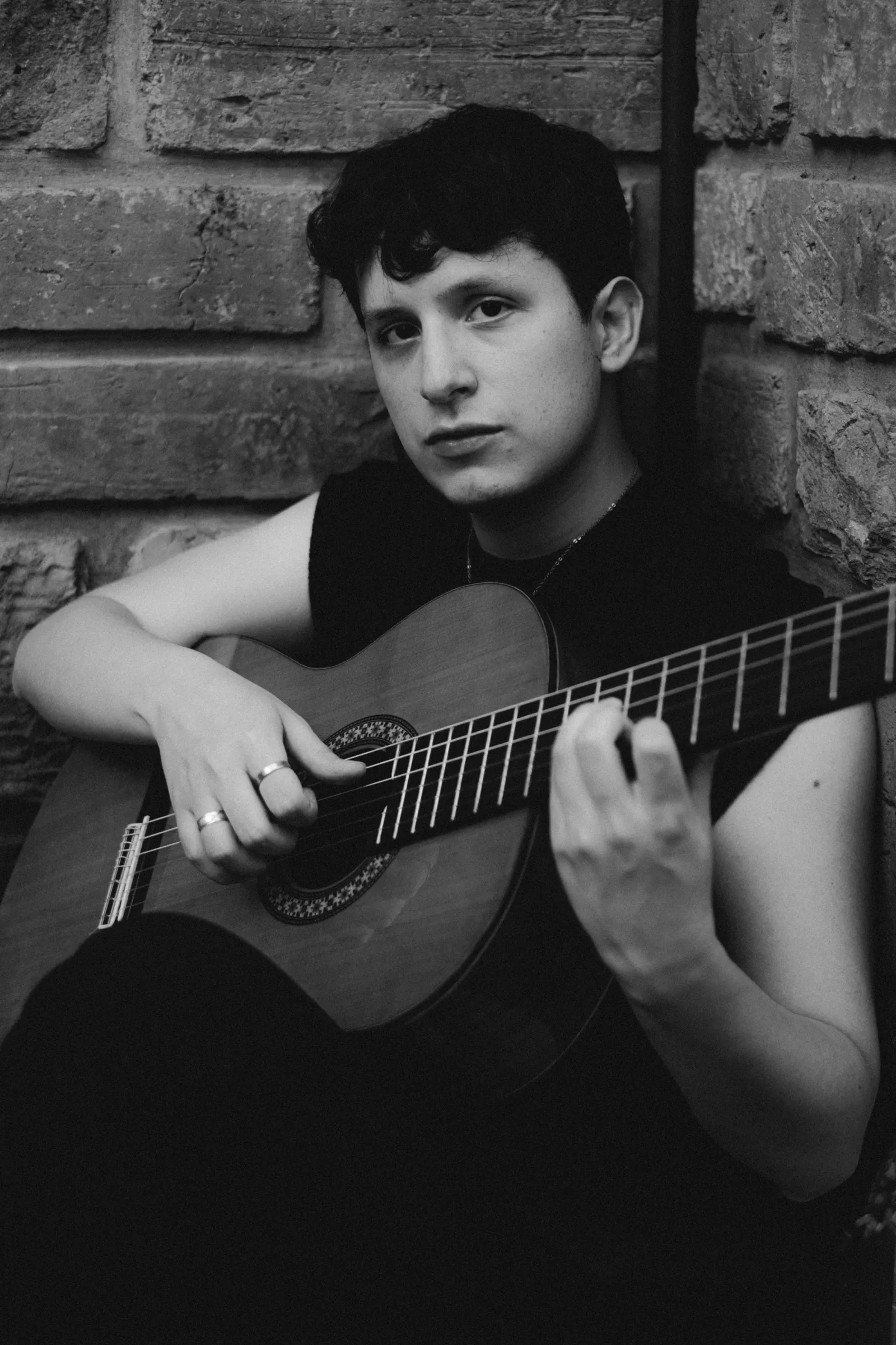 Chéri sentado contra un muro de piedra, tocando la guitarra, en blanco y negro
